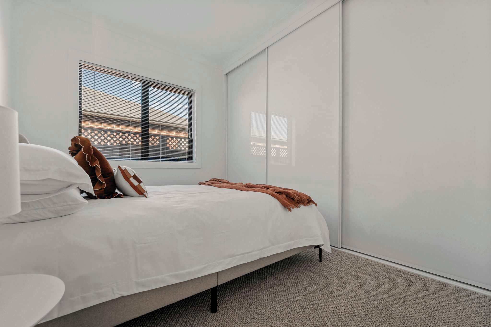 A bedroom with a bed, a window with blinds, a white wardrobe, and a carpeted floor.