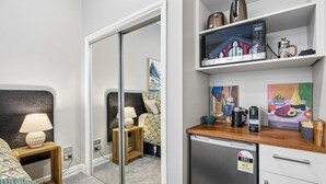 Ett modernt hotellrum med en säng, en sänglampa, ett litet träbord, en spegel, ett litet kök med kylskåp, en kaffebryggare och en mikrovågsugn.