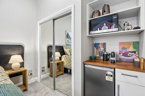 A modern hotel room with a bed, bedside lamp, a small wooden nightstand, a mirror, a kitchenette with a refrigerator, a coffee maker, and a microwave. - Chapel on Moray - studio room with bathroom and kitchenette (Dunedin)