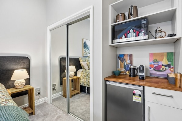 A modern hotel room with a bed, bedside lamp, a small wooden nightstand, a mirror, a kitchenette with a refrigerator, a coffee maker, and a microwave.