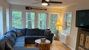 A living room with a sectional sofa, a round coffee table, a ceiling fan, a TV, and large windows with a view of trees. - Fun 5 Bedroom Lakefront Cottage (Bethel)