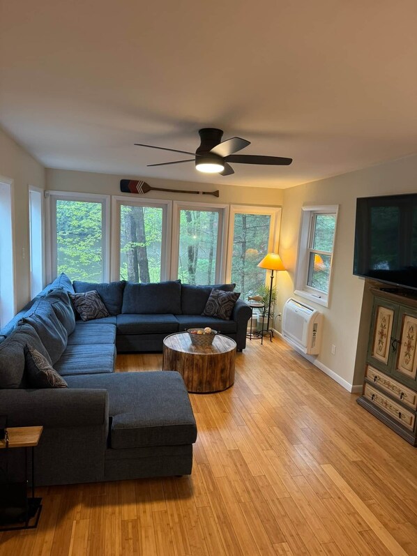 A living room with a sectional sofa, a round coffee table, a ceiling fan, a TV, and large windows with a view of trees. - Fun 5 Bedroom Lakefront Cottage (Bethel)
