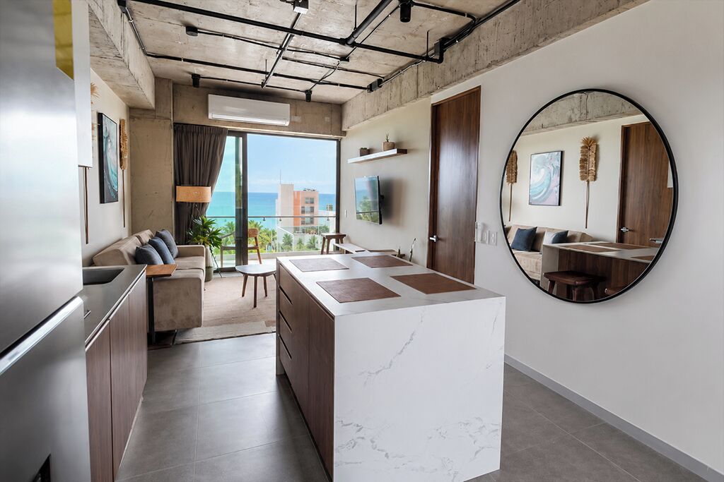 A modern kitchen with a marble island, a dining area with a round mirror, and a view of the sea.