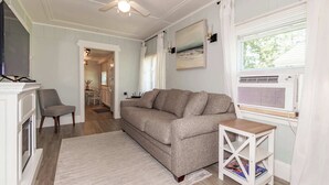 Un salon comprenant un canapé, un téléviseur, un ventilateur de plafond, une cheminée et une fenêtre avec des stores.