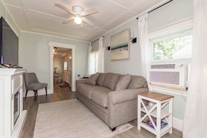 Ein Wohnzimmer mit Sofa, Fernseher, Deckenventilator, Kamin und einem Fenster mit Jalousien.