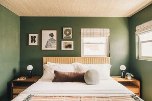 Ein Schlafzimmer mit Bett, Nachttischen, einem Fenster mit Vorhängen und Wandbildern.