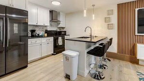 Une cuisine moderne avec un réfrigérateur en acier inoxydable, des armoires blanches, un plan de travail noir, un évier et un tabouret de bar.