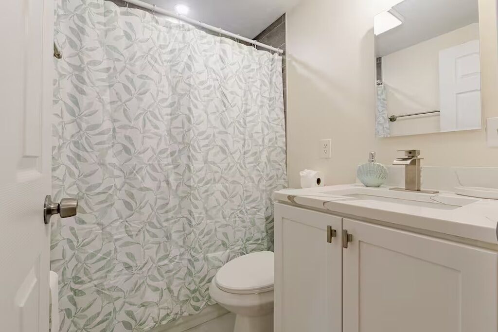 Une salle de bain comprenant des toilettes, un lavabo avec un miroir et un rideau de douche orné d’un motif de feuilles.