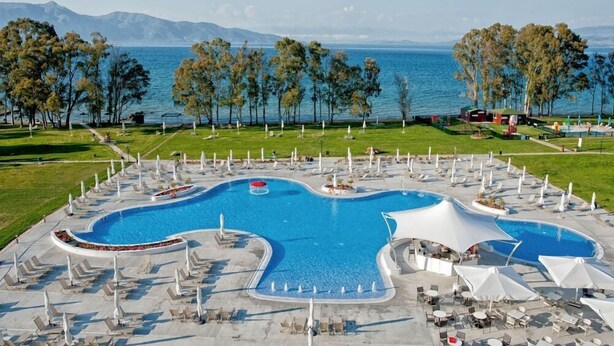 Una gran piscina al aire libre con varias secciones, rodeada de sillones de descanso y sombrillas, con vistas al mar y a las montañas al fondo.