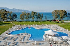 Una gran piscina al aire libre con varias secciones, rodeada de sillones de descanso y sombrillas, con vistas al mar y a las montañas al fondo.