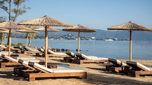 Una playa con sillas de madera, sombrillas de paja y una vista despejada del mar, con colinas a lo lejos.
