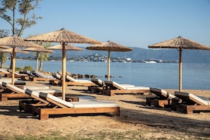 Una spiaggia con lettini di legno e ombrelloni di paglia, vista libera sul mare e colline sullo sfondo.