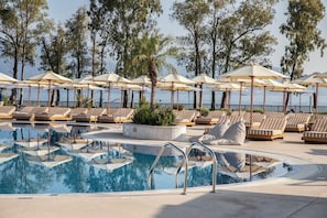 Ein Poolbereich mit Liegestühlen und Sonnenschirmen, umgeben von Bäumen und Blick aufs Meer.