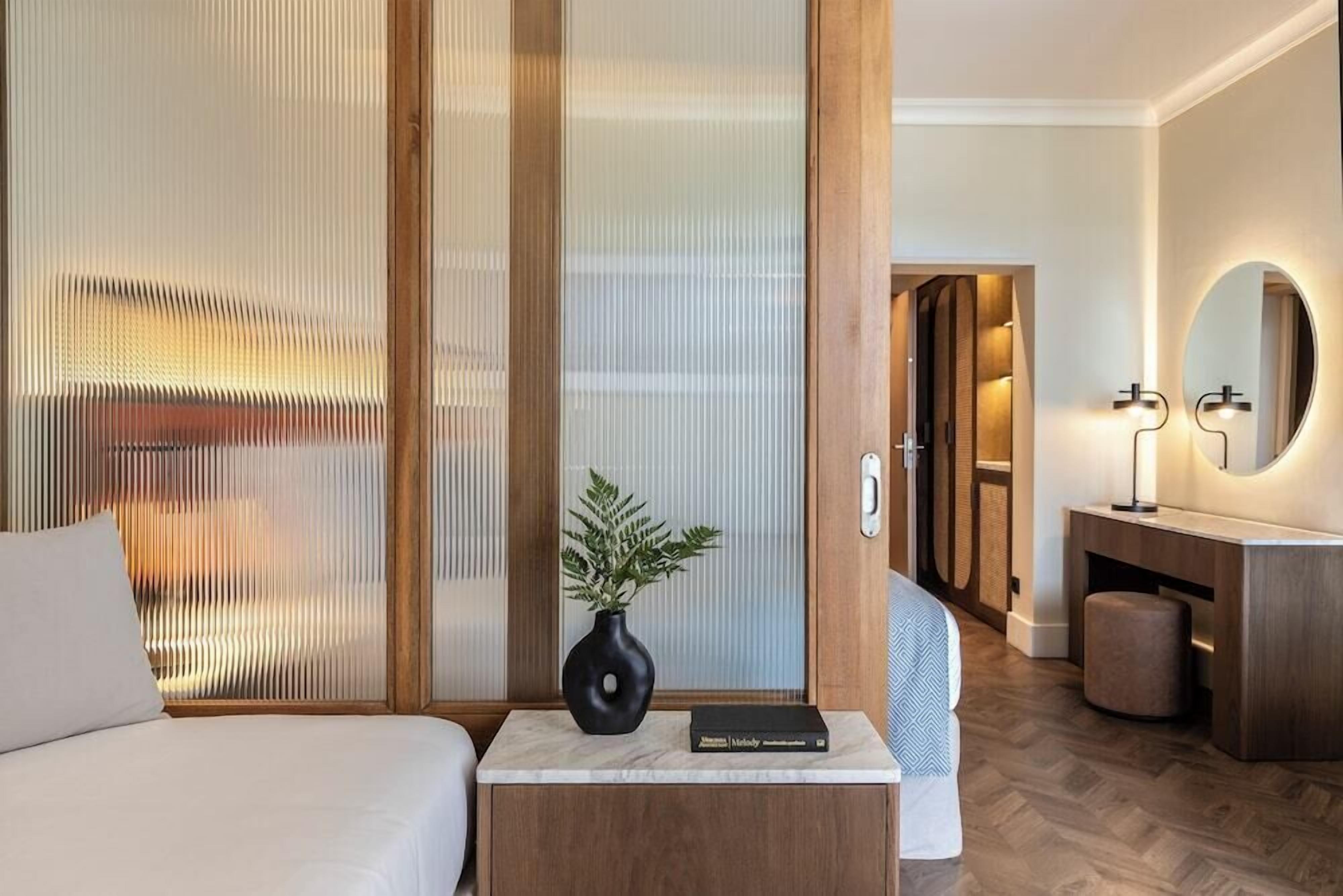 Una habitación de hotel con cama, una mesita de noche con un libro, un jarrón con una planta, un espejo y suelo de madera.
