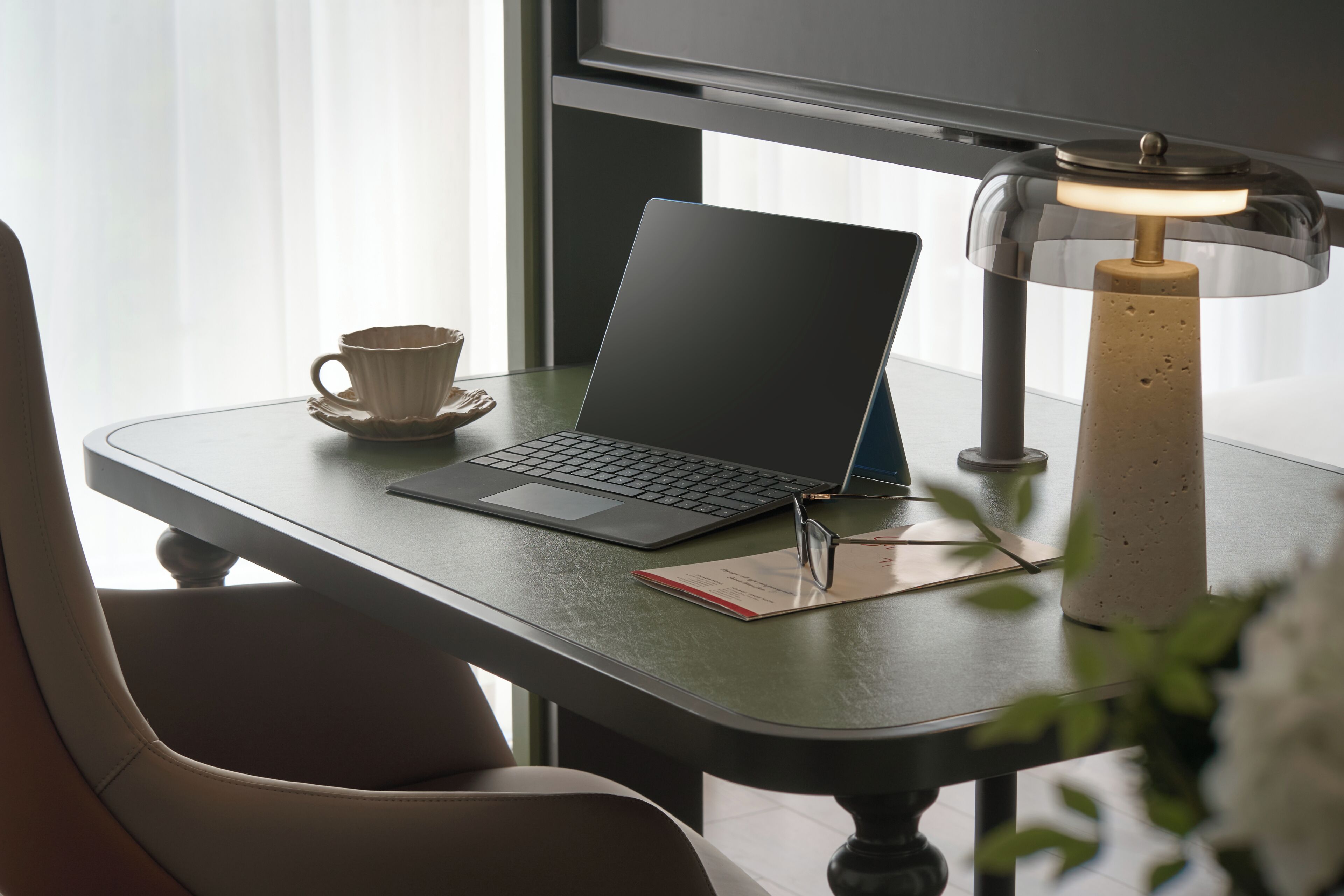 A laptop on a table with a lamp, a cup, and a notebook.