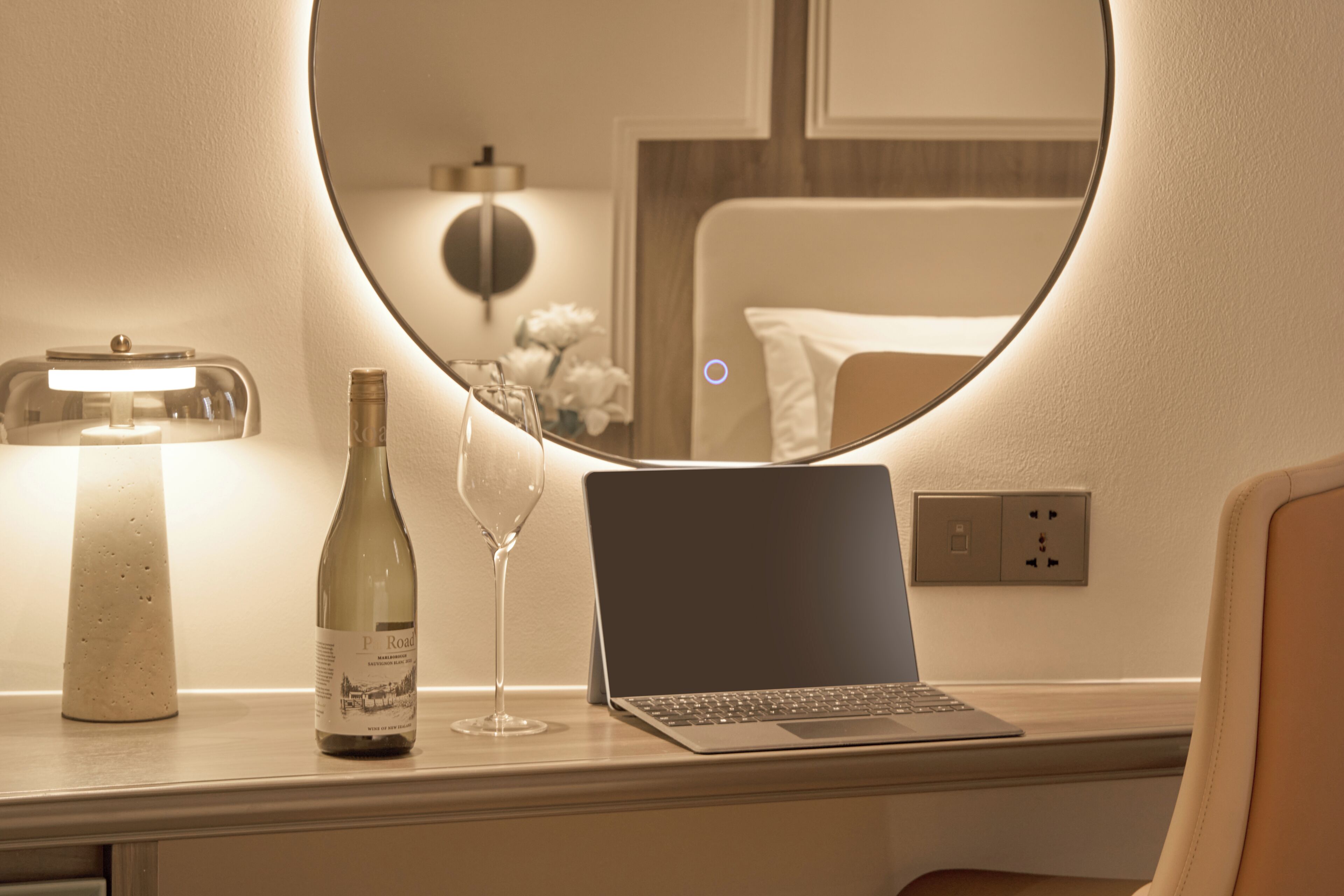 A laptop on a desk with a wine bottle and glass, a round mirror, and a lamp.