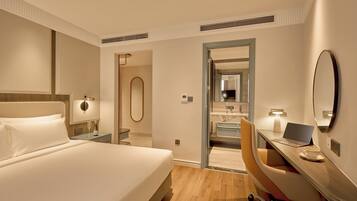 Habitación de hotel con cama, escritorio, silla y baño visible a través de una puerta abierta.