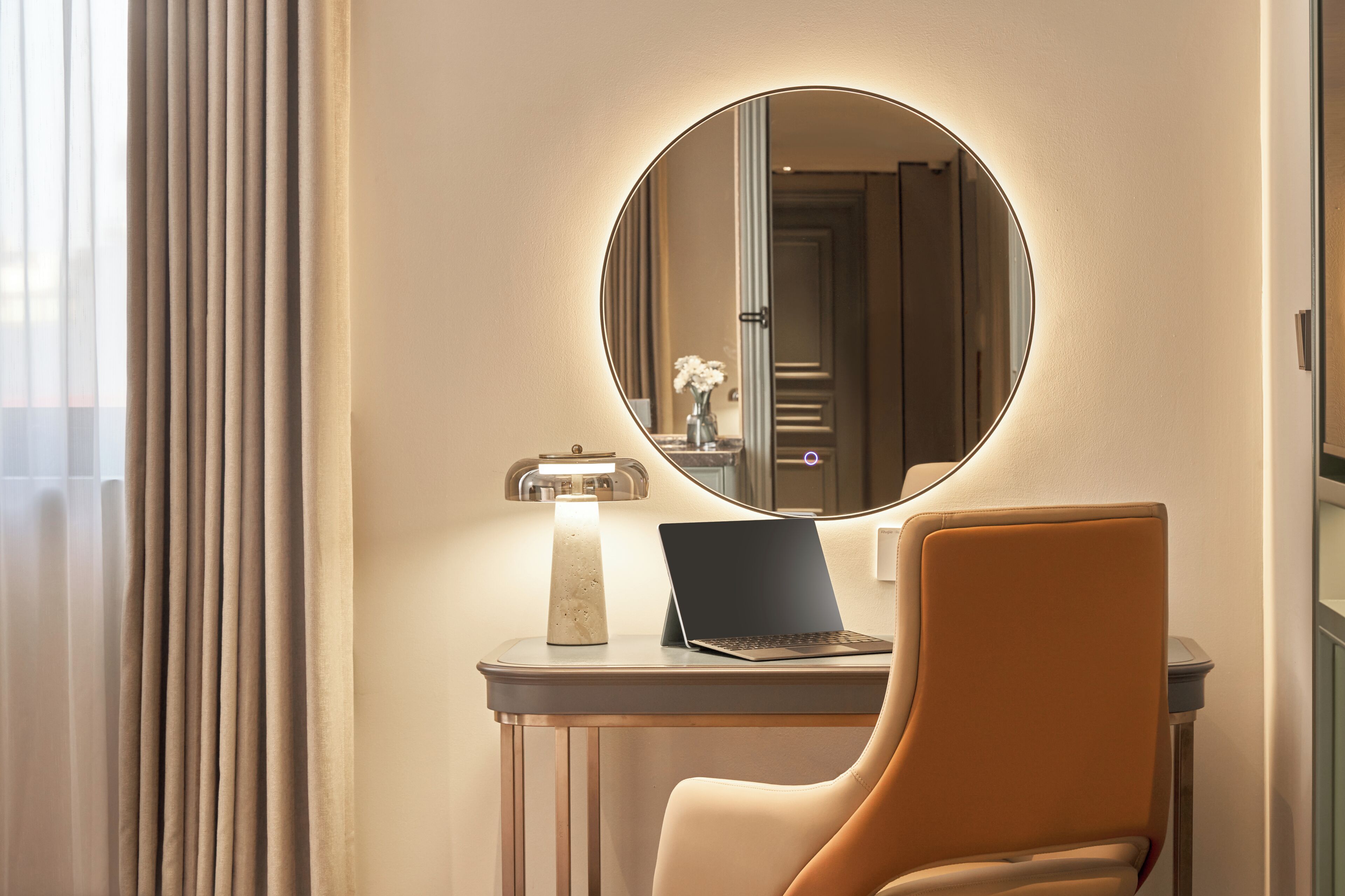 A room with a round mirror, a laptop on a small table, a chair, and a lamp.