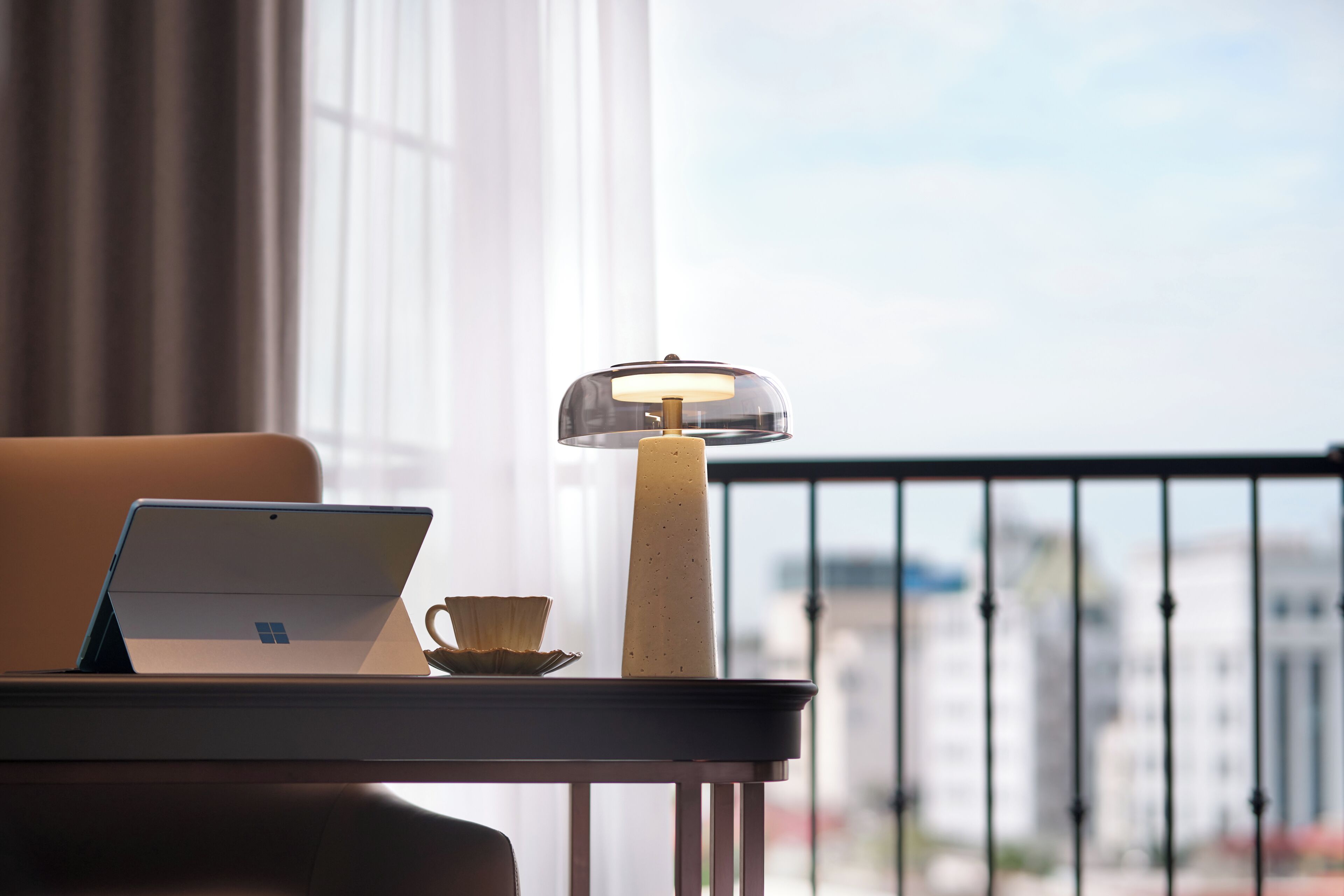 A laptop on a table with a candle and a cup, near a window with a view of buildings.