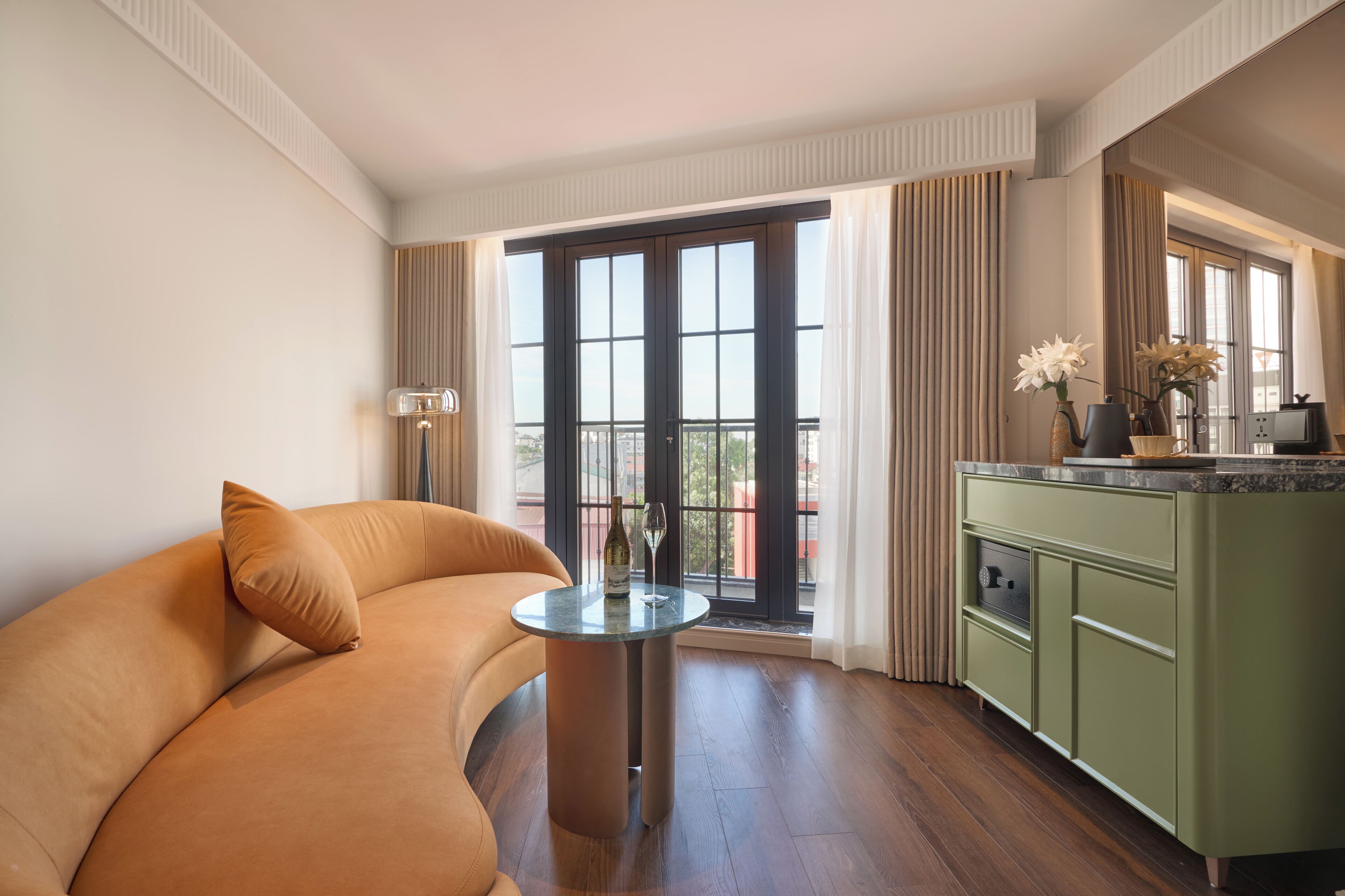 A modern hotel room with a curved sofa, a small round table, a green minibar, and a large sliding glass door leading to a balcony.
