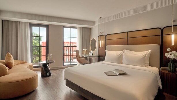 A hotel room with a large bed, a sofa, a small table with a chair, a mirror, and a vase with flowers. - Solaria d'Oriente Hotel (Hanoi)