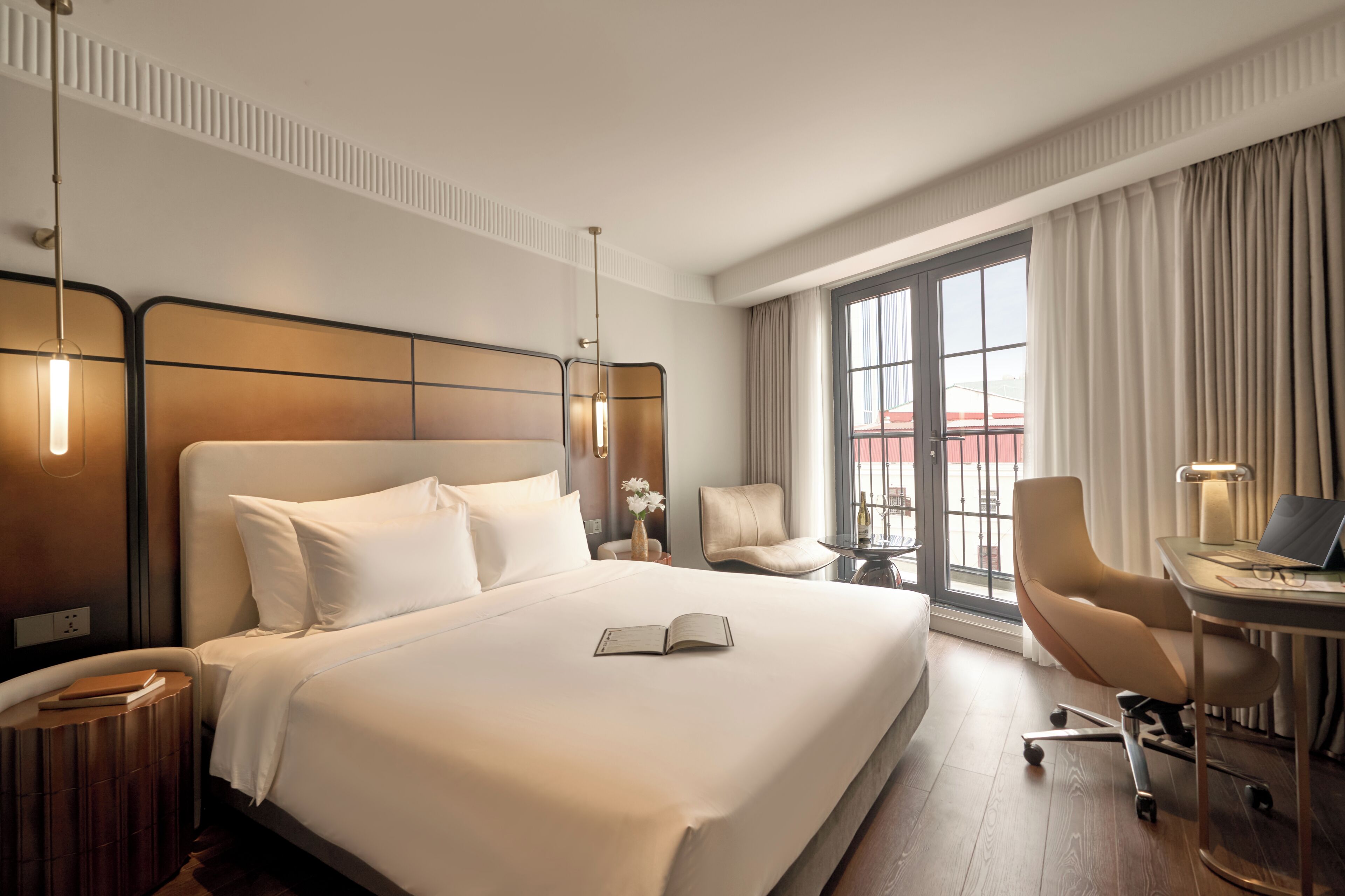 A hotel room with a large bed, a desk with a laptop, a chair, and a view of the outside through a window.