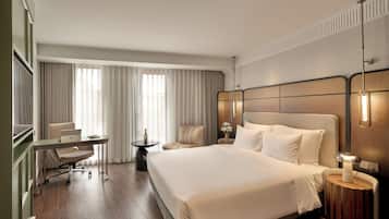 Quarto de hotel com uma cama grande, uma escrivaninha com um laptop, uma cadeira, uma mesinha redonda com uma garrafa e uma mesa de cabeceira com uma luminária.
