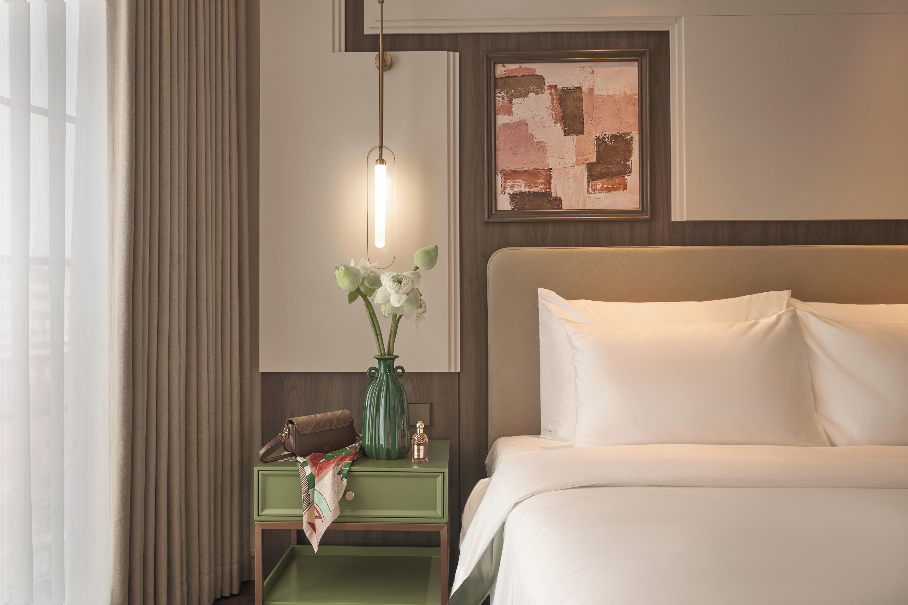 A hotel room with a bed, a nightstand with a vase of flowers, a lamp, and a painting on the wall.