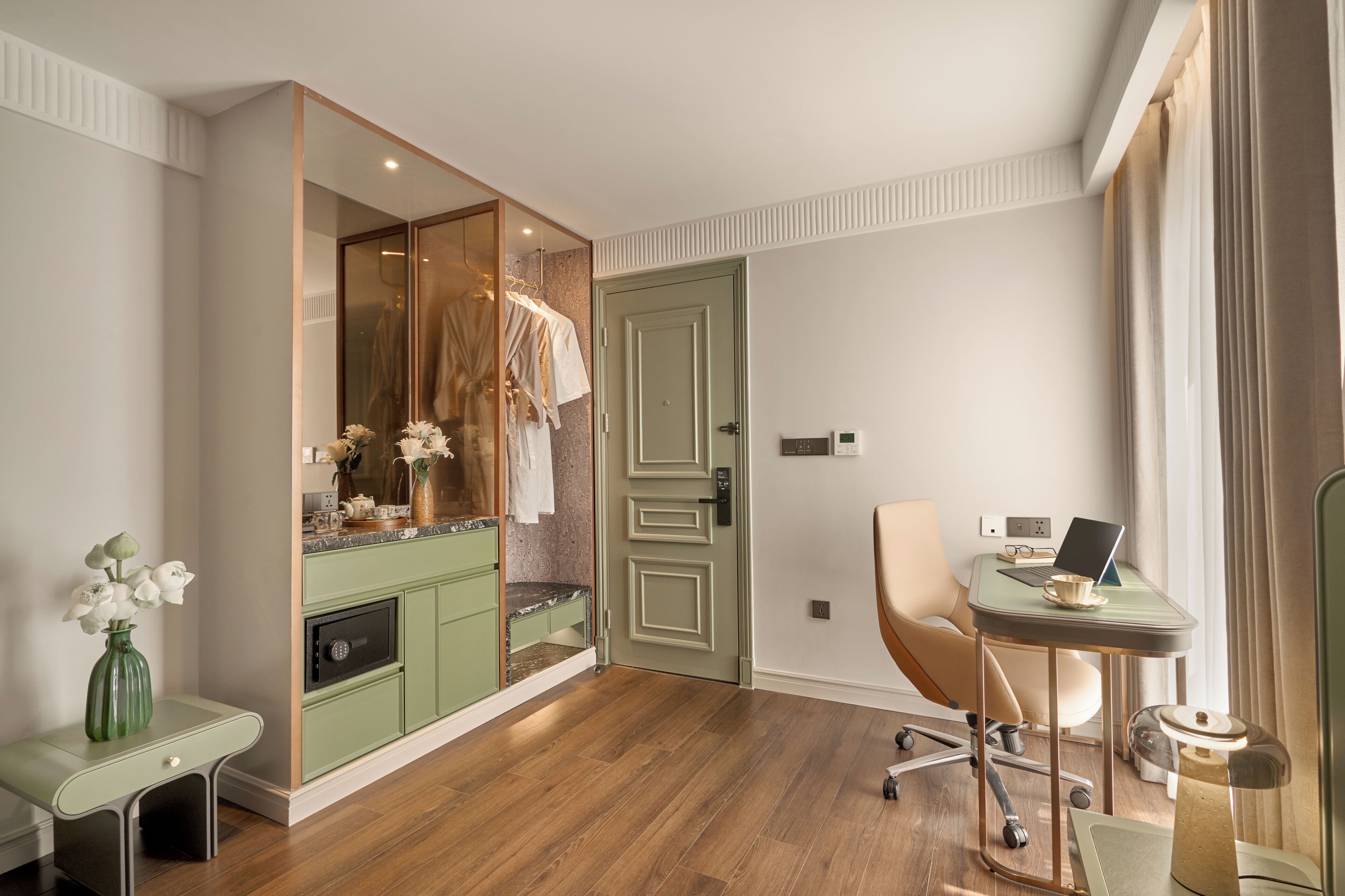Habitación de hotel con un tocador verde, un espejo, un escritorio con una laptop, una silla y un florero con flores.