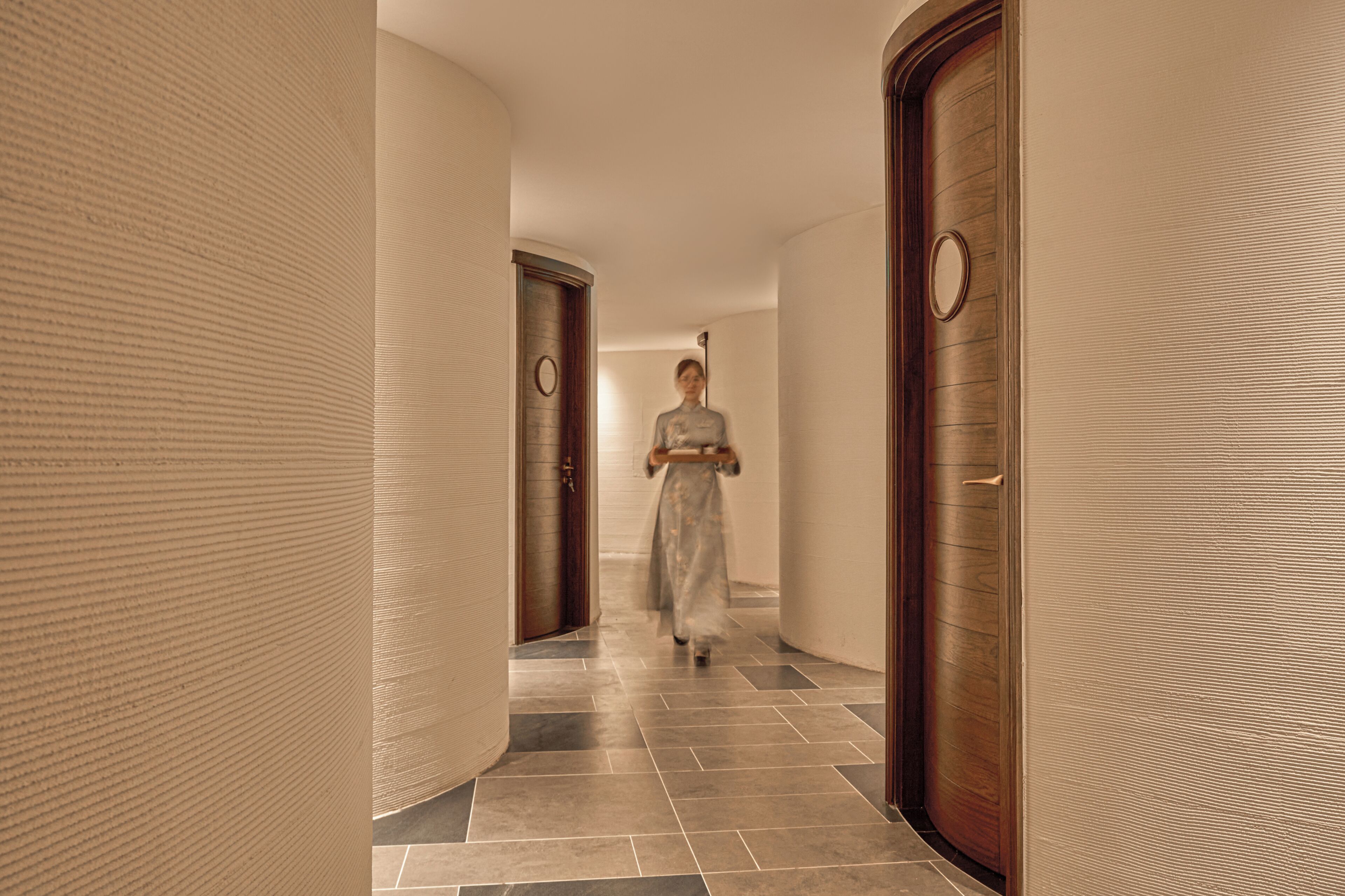 A corridor with a person walking, wooden doors, and a patterned wall.