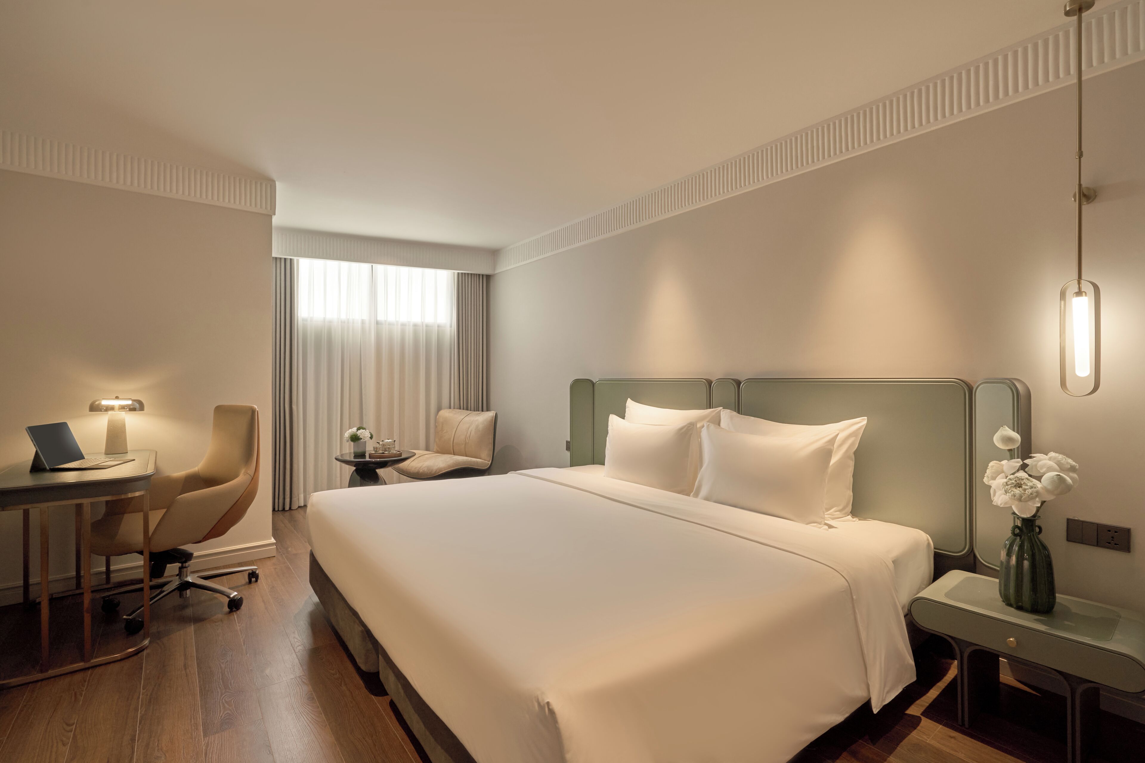 A hotel room with a large bed, a desk with a laptop, a chair, a small table with a vase, and a nightstand.