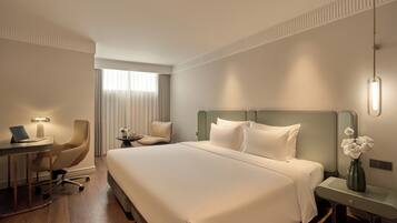 A hotel room with a large bed, a desk with a laptop, a chair, a small table with a vase, and a nightstand.