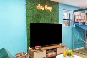 Una habitación con una televisión de pantalla plana sobre un soporte de madera, un letrero de neón que dice 'Stay Cozy', una pared verde y un aro de baloncesto.