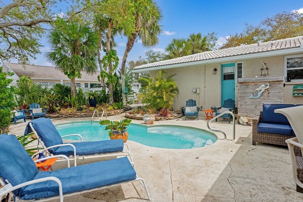 A pool area with lounge chairs, a small house with a tiled roof, and various plants.