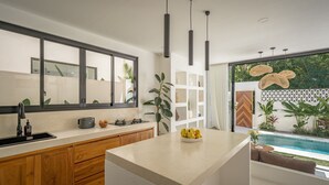 A modern kitchen with a large island, wooden cabinets, a built-in oven, and a view of a pool. - Villa Nikki Uluwatu (Uluwatu)