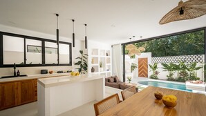 A modern kitchen with a central island, wooden dining table, and a view of a swimming pool. - Villa Nikki Uluwatu (Uluwatu)