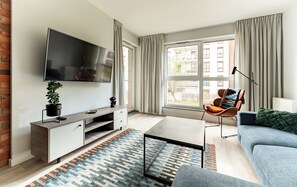 Ein modernes Wohnzimmer mit Flachbildfernseher, einer Couch, einem Couchtisch und einem großen Fenster mit Vorhängen.