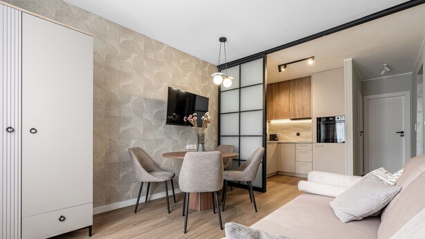 A modern living room with a sofa, a dining table with chairs, a TV mounted on the wall, and a kitchen area with cabinets and appliances. - Przymorze Kaszmirowy Apartment (Gdańsk)
