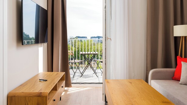 A modern hotel room with a TV, wooden furniture, a balcony with outdoor seating, and a view of the city. - Warszawa Rakowiec Deluxe Apartment (Warszawa)