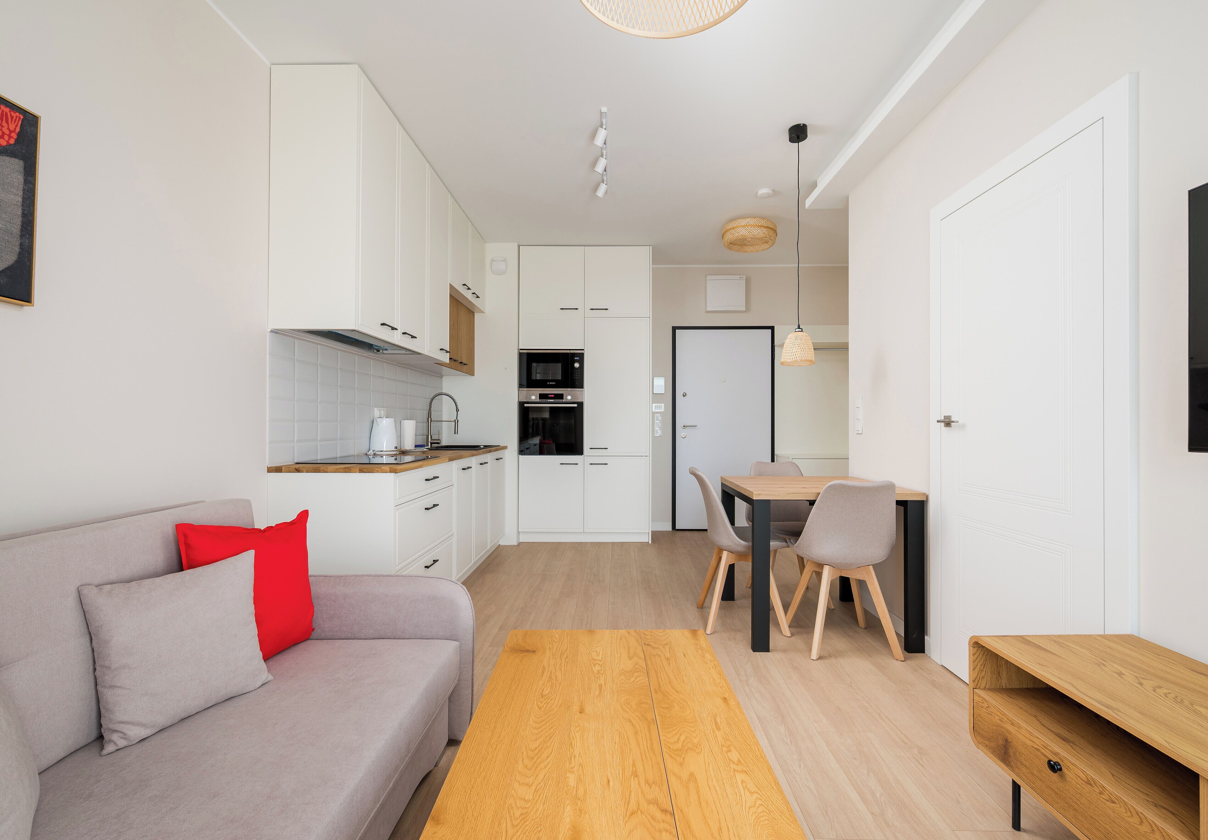 A modern kitchen with white cabinets, a dining area with a wooden table and chairs, and a sofa with red and grey cushions.