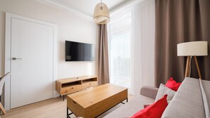 A modern living room with a wooden coffee table, a sofa with red cushions, a TV mounted on the wall, and a large window with curtains. - Warszawa Rakowiec Deluxe Apartment (Warszawa)
