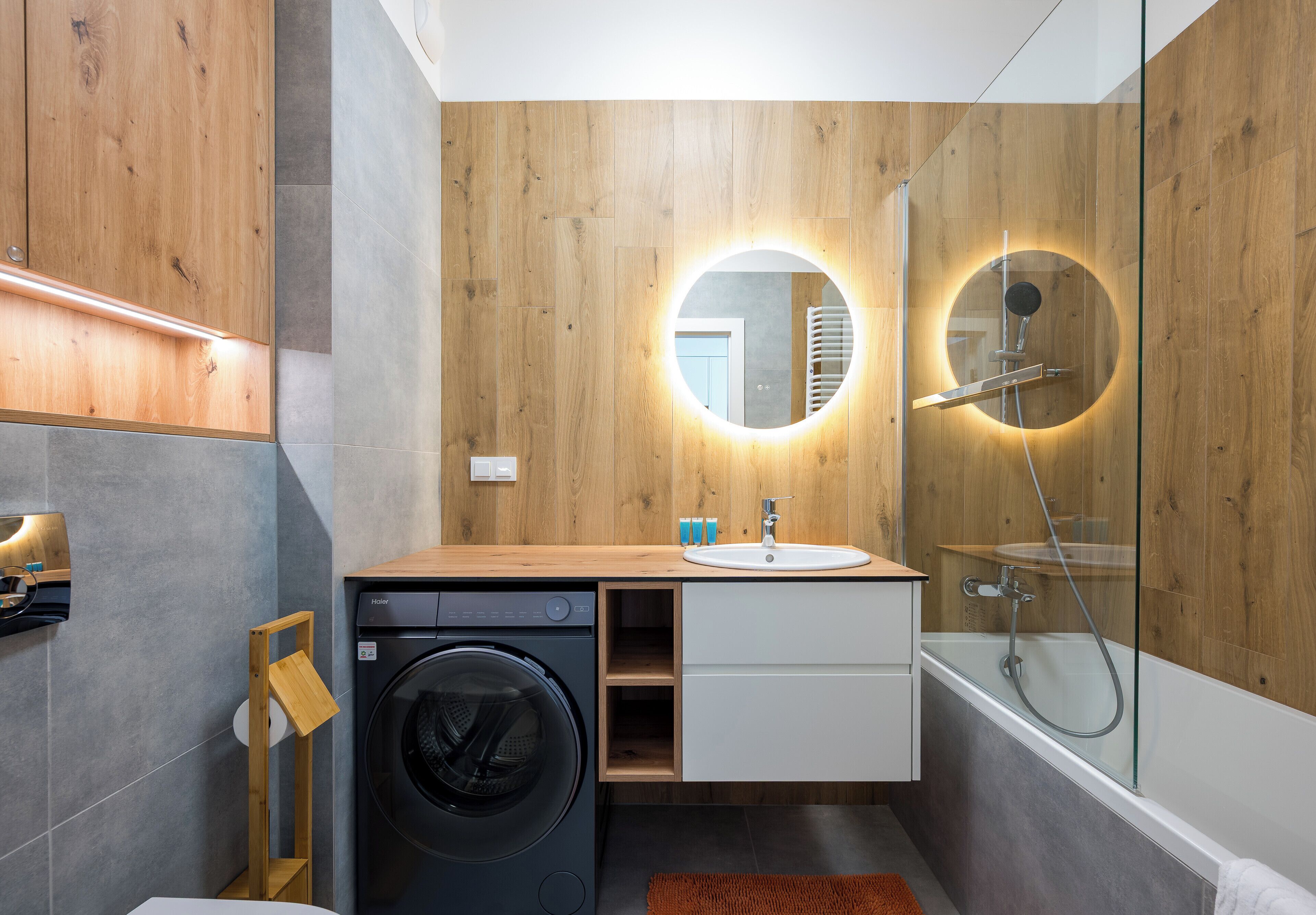 Un baño moderno con tocador de madera, lavadora, cabina de ducha de vidrio y espejo redondo con luz.