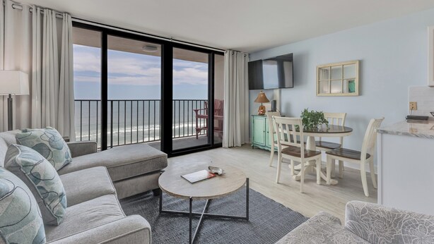 En moderne stue med en sofa, et rundt sofabord, et spiseområde med bord og stole, et fjernsyn monteret på væggen og en balkon med havudsigt.