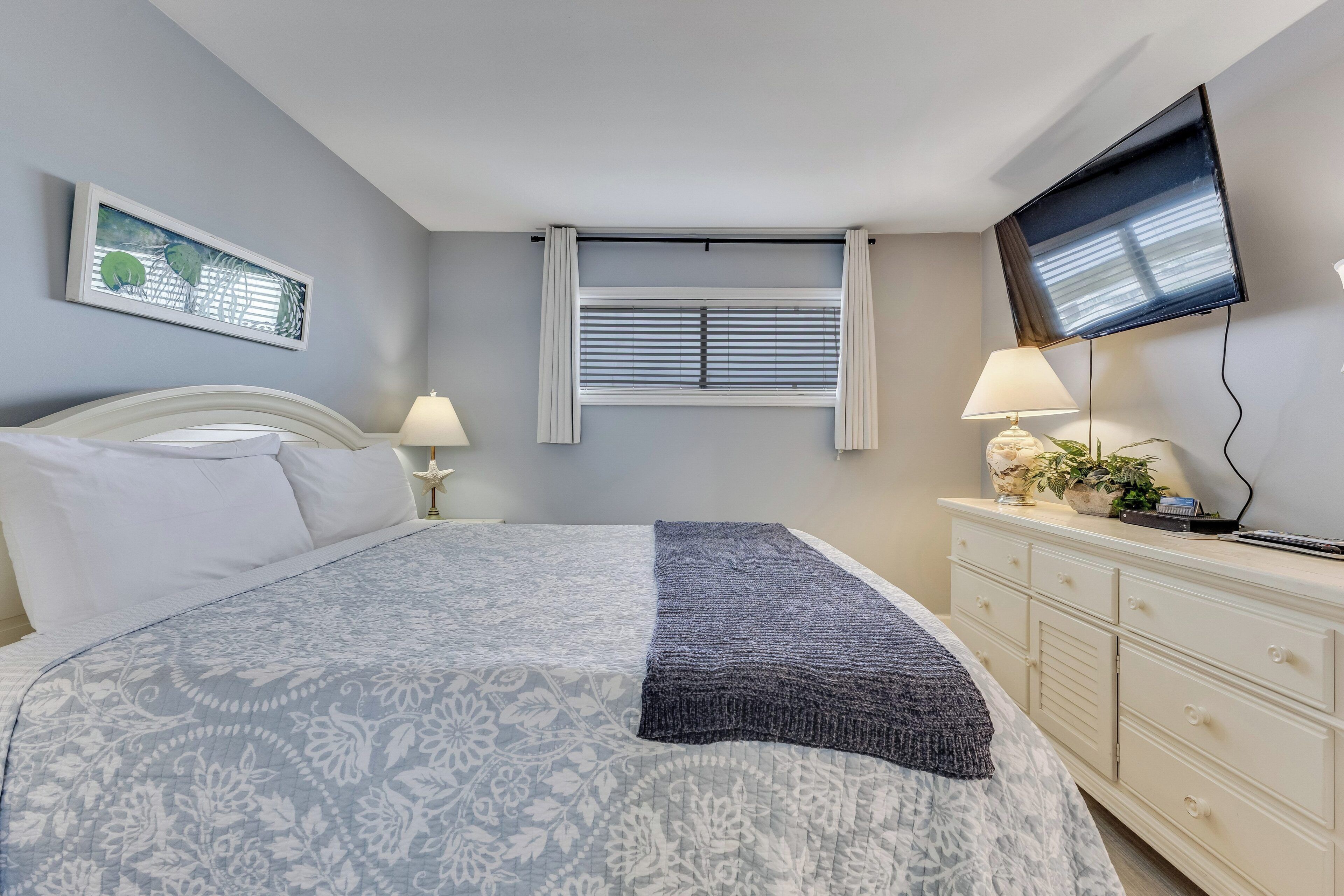 Een slaapkamer met een bed, nachtkastjes, een dressoir, een televisie, een raam met jaloezieën en een schilderij aan de muur.