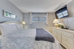Ein Schlafzimmer mit Bett, Nachttischen, Kommode, Fernseher, Fenster mit Jalousien und einem Gemälde an der Wand.