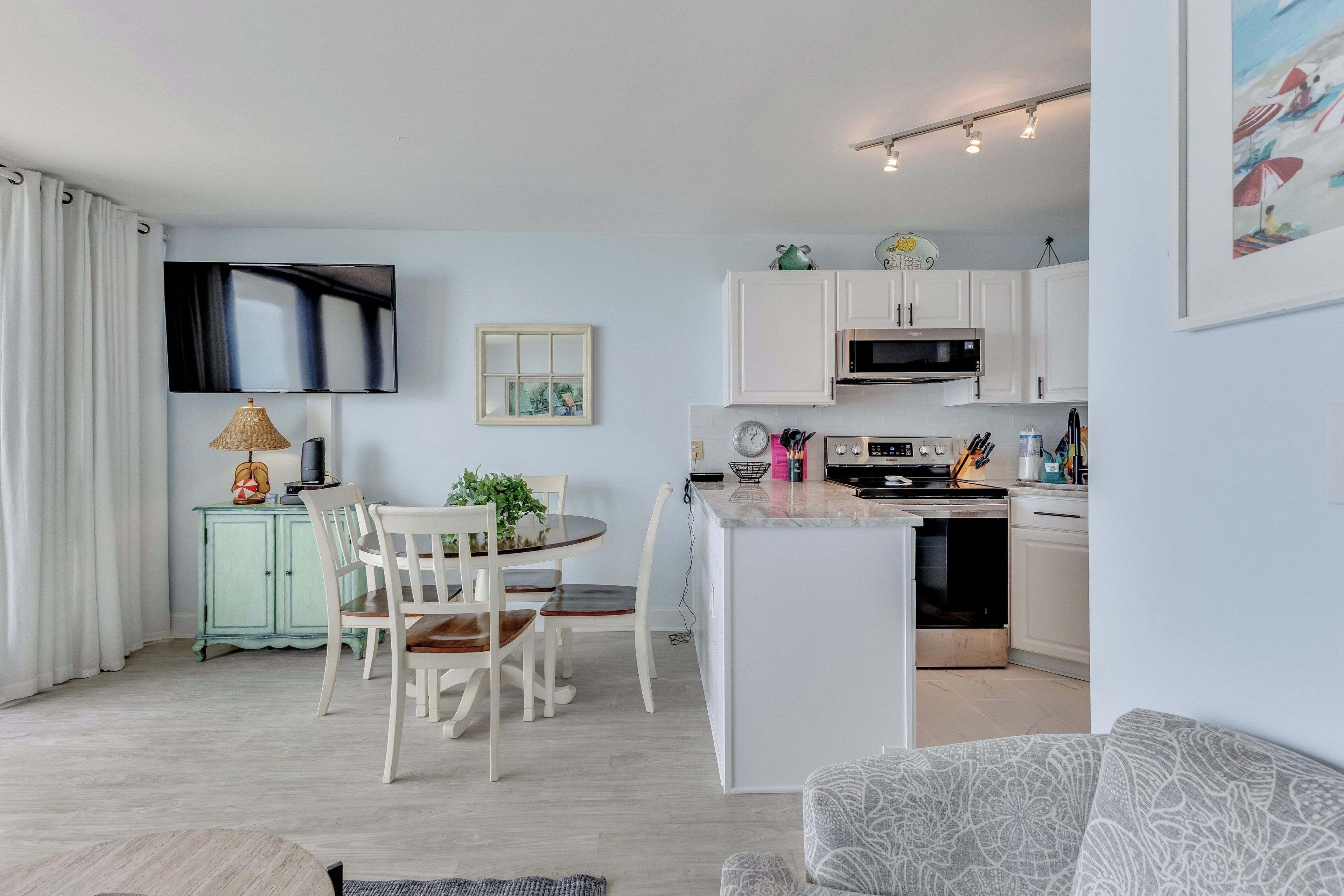 Een moderne keuken met witte kasten, een eethoek met tafel en stoelen, een aan de muur bevestigde televisie en een klein kookeiland met een magnetron.
