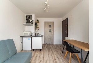 A compact kitchenette with a microwave, coffee maker, and a small refrigerator. There is a wooden dining table with a black chair, a grey sofa, and a door leading to another room. - Gdynia Morska Horizon Studio (Gdynia)