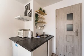 A compact kitchenette with a microwave, toaster, and sink. There are wooden shelves with plants and a door with a frosted glass panel. - Gdynia Morska Horizon Studio (Gdynia)