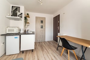 Een kleine keuken met een magnetron, koffiezetapparaat en een kleine koelkast. Er staat een houten tafel met een zwarte stoel bij, en op de achtergrond een houten deur.