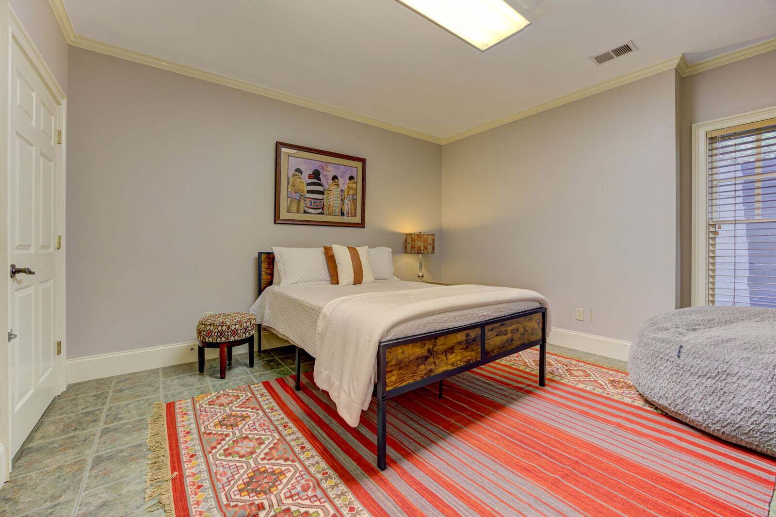 Un dormitorio con cama, mesita de noche, silla, alfombra, cuadro y ventana.